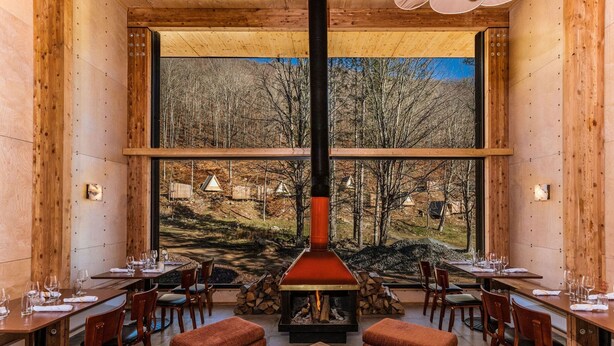 A cozy dining area with wooden walls, large windows, and a central wood-burning stove. Tables are set with glasses and cutlery. - Eastwind Oliverea Valley (Big Indian)