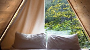 A triangular wooden tent with a bed inside, featuring a view of a forest with yellow foliage. - Eastwind Oliverea Valley (Big Indian)