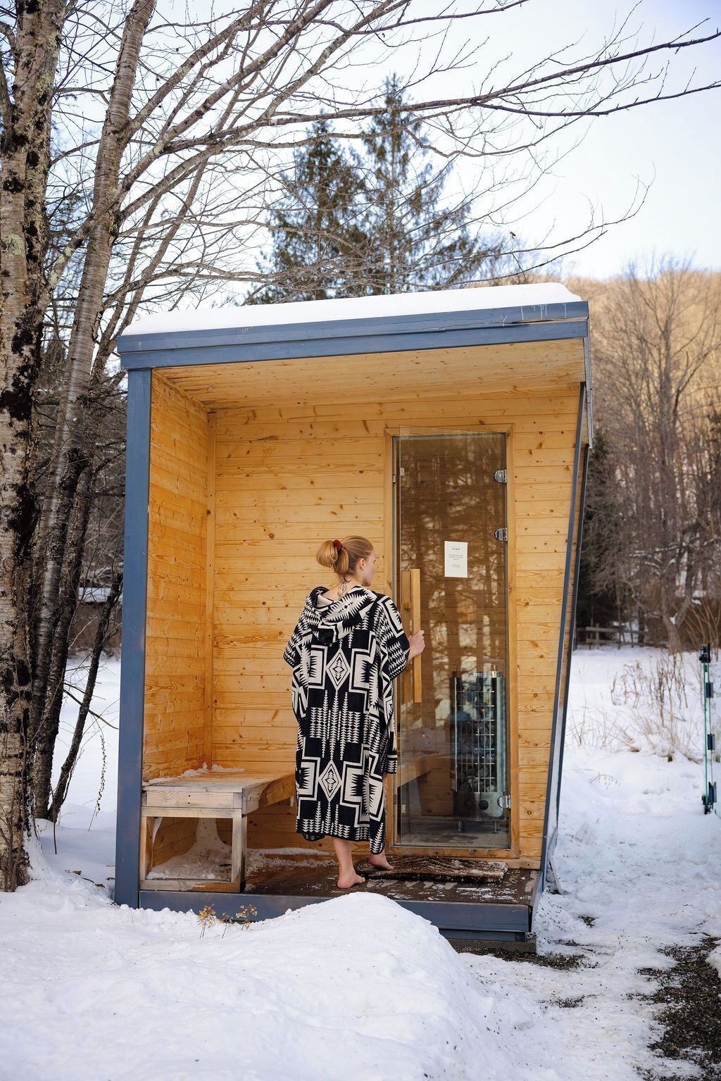 A small wooden cabin with a glass door, a bench outside, and a snowy ground.