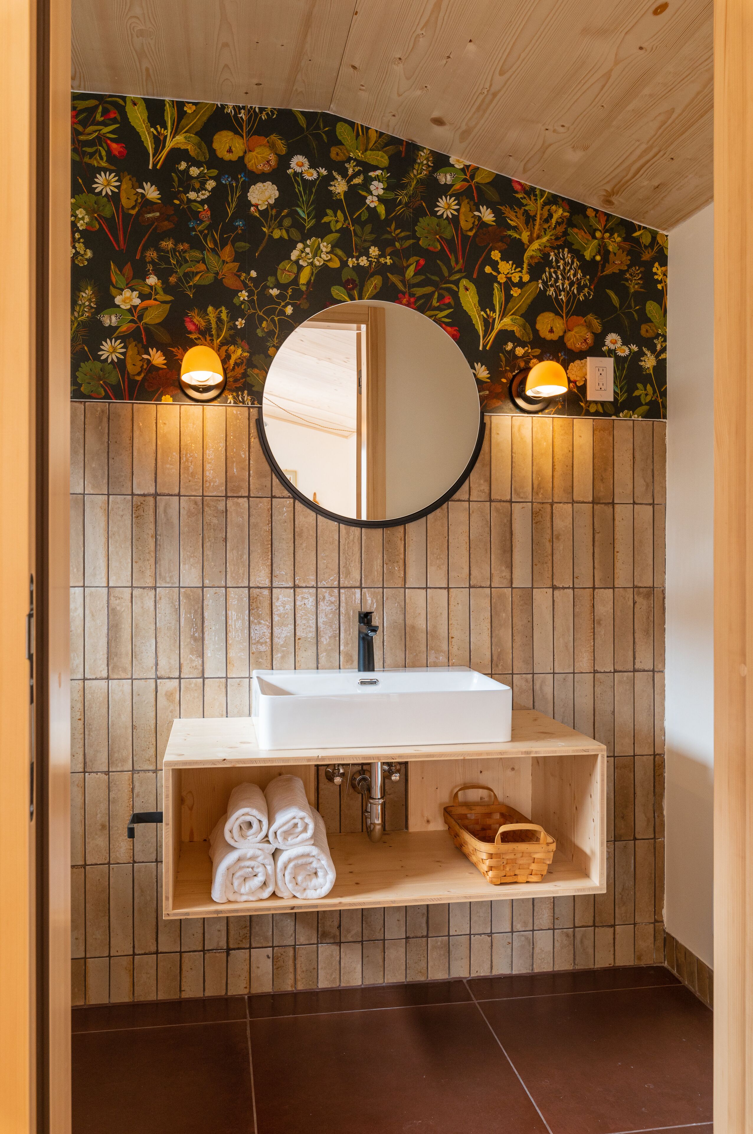A bathroom with a wooden vanity, a round mirror, and a floral wallpaper design.