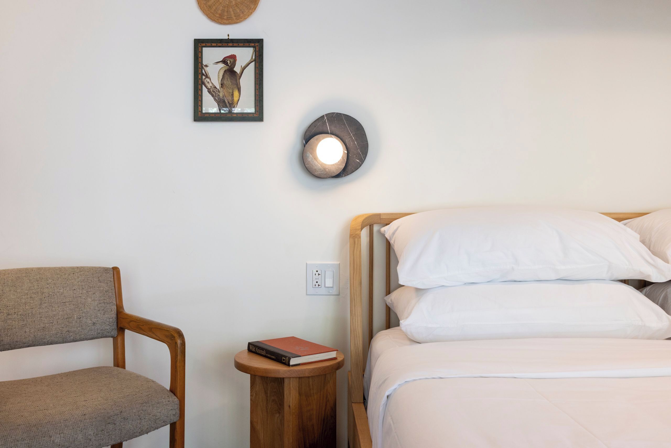 Un dormitorio ordenado con una cama de madera, una pequeña mesa redonda con un libro, una silla, una lámpara de pared y un cuadro enmarcado.