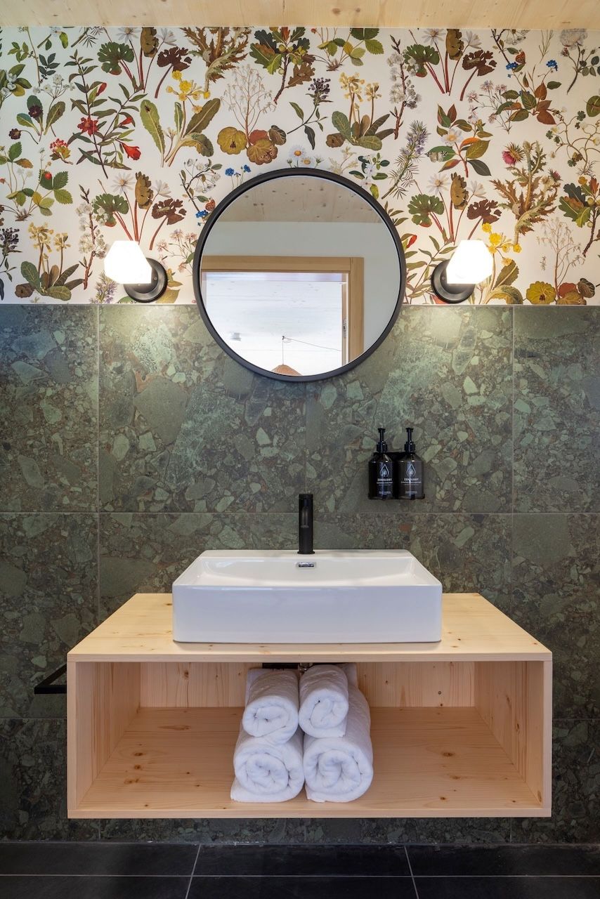 A bathroom with a white sink, a wooden shelf with rolled towels, a mirror, and a floral wallpaper.