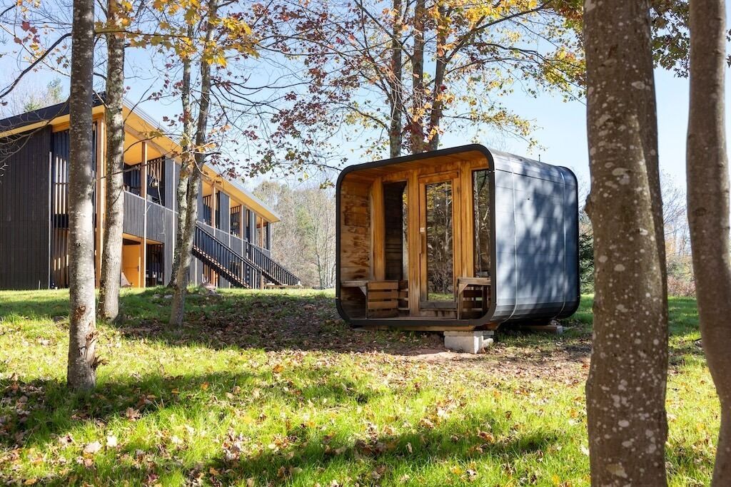 A modern wooden cabin with a glass enclosure, surrounded by trees and a grassy area.