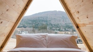 A triangular wooden room with a bed and pillows, a window, and a view of a forested hill. - Eastwind Windham (Windham)