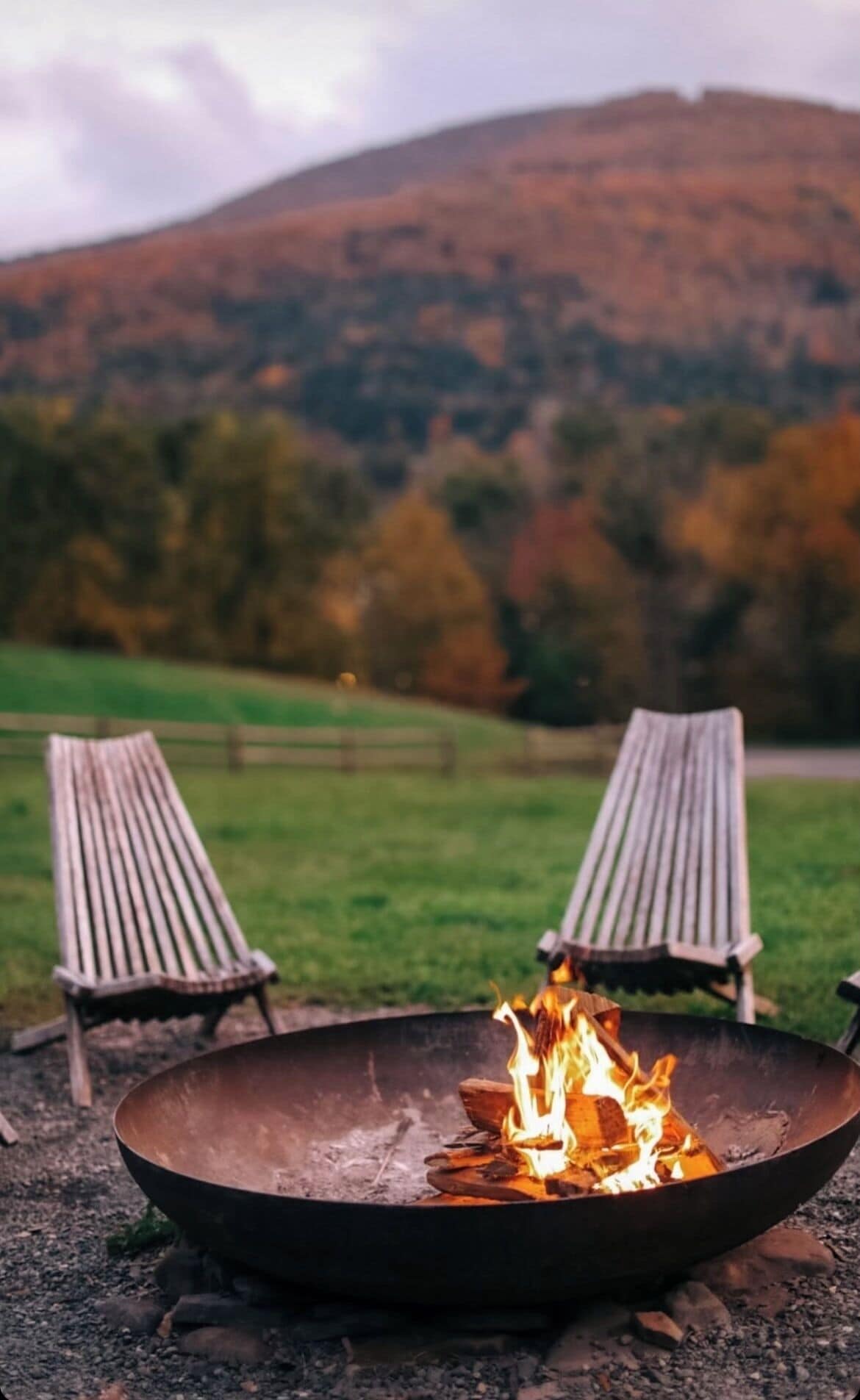 A campfire with two wooden chairs facing it, set against a backdrop of a grassy field and a forested hill.