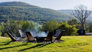 A campfire with four chairs arranged around it in a grassy field with a forested hill in the background.