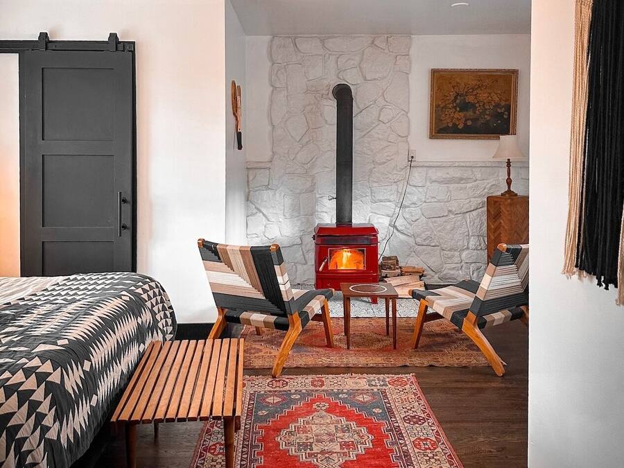 Una habitación acogedora con una cama, un banco de madera, un estufa de leña roja y una alfombra estampada.