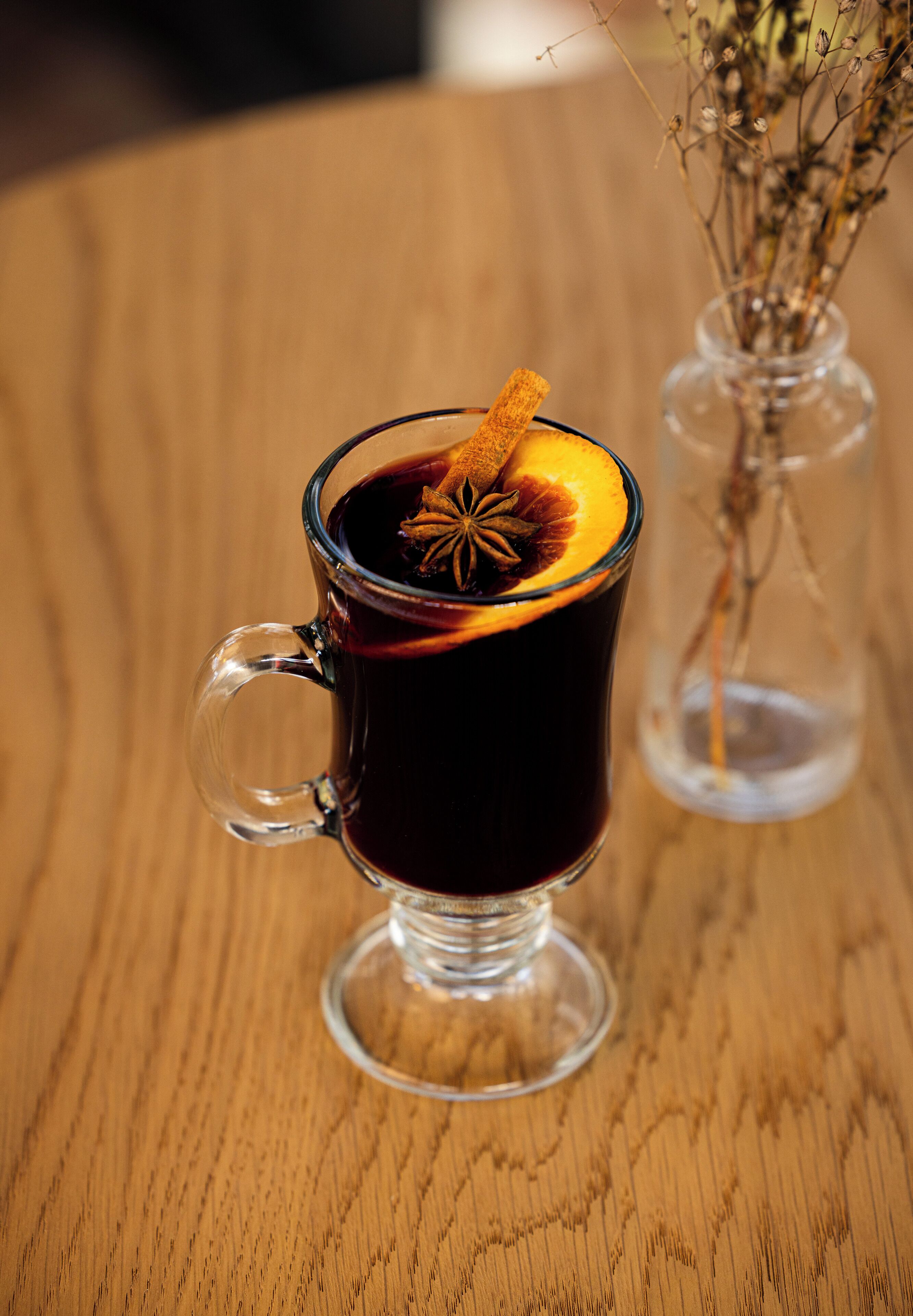 A clear glass mug with a handle, filled with a dark liquid, possibly tea or coffee, and garnished with a cinnamon stick and a slice of orange.