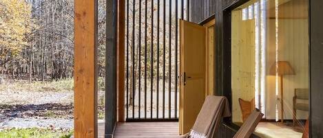 A wooden deck with a chair and a table, a large window, and a door leading to a room.