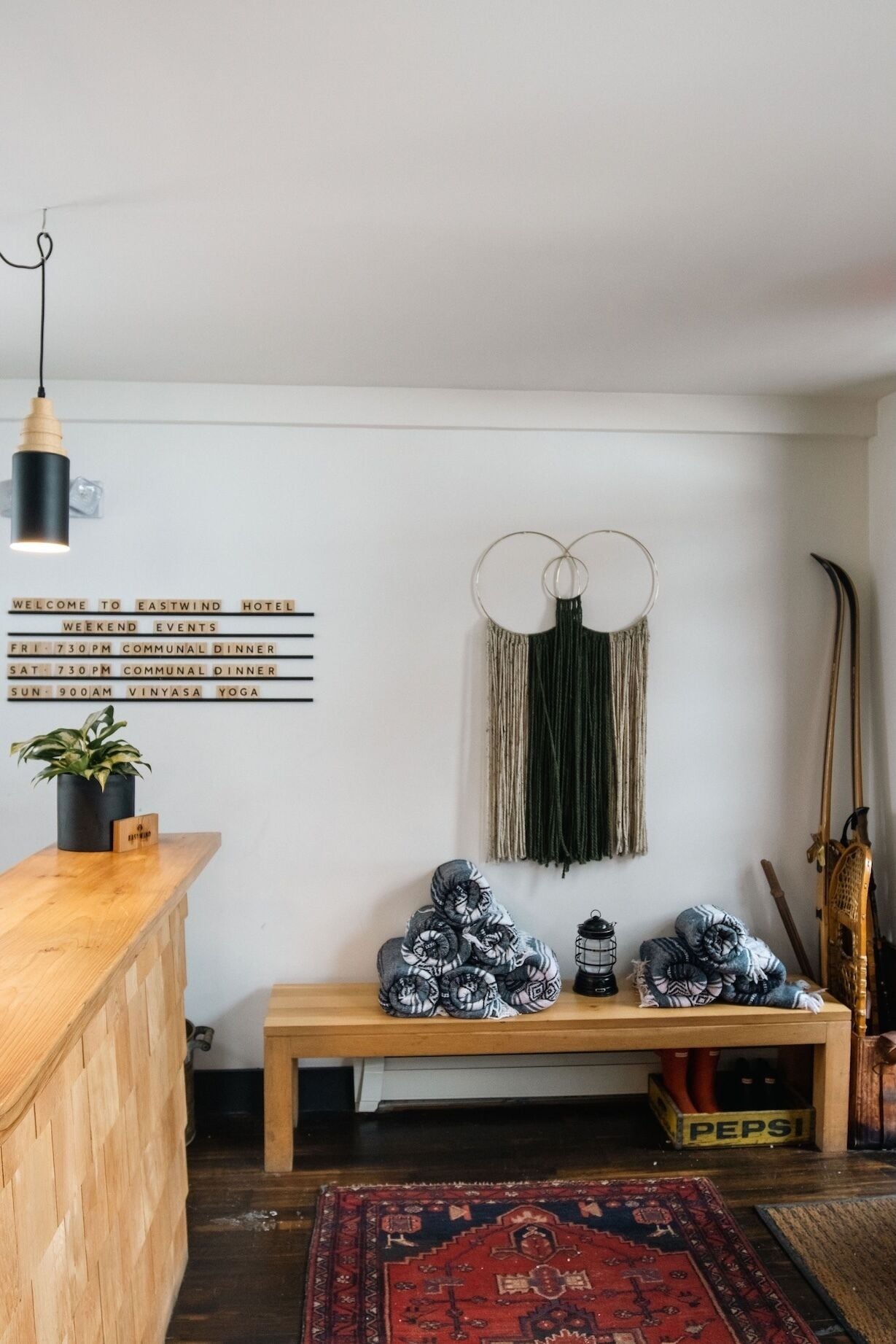 A hotel reception area with a wooden counter, a black pendant light, a wall-mounted sign listing weekend events, a bench with rolled-up blankets, a macrame wall hanging, and a vintage vacuum cleaner.