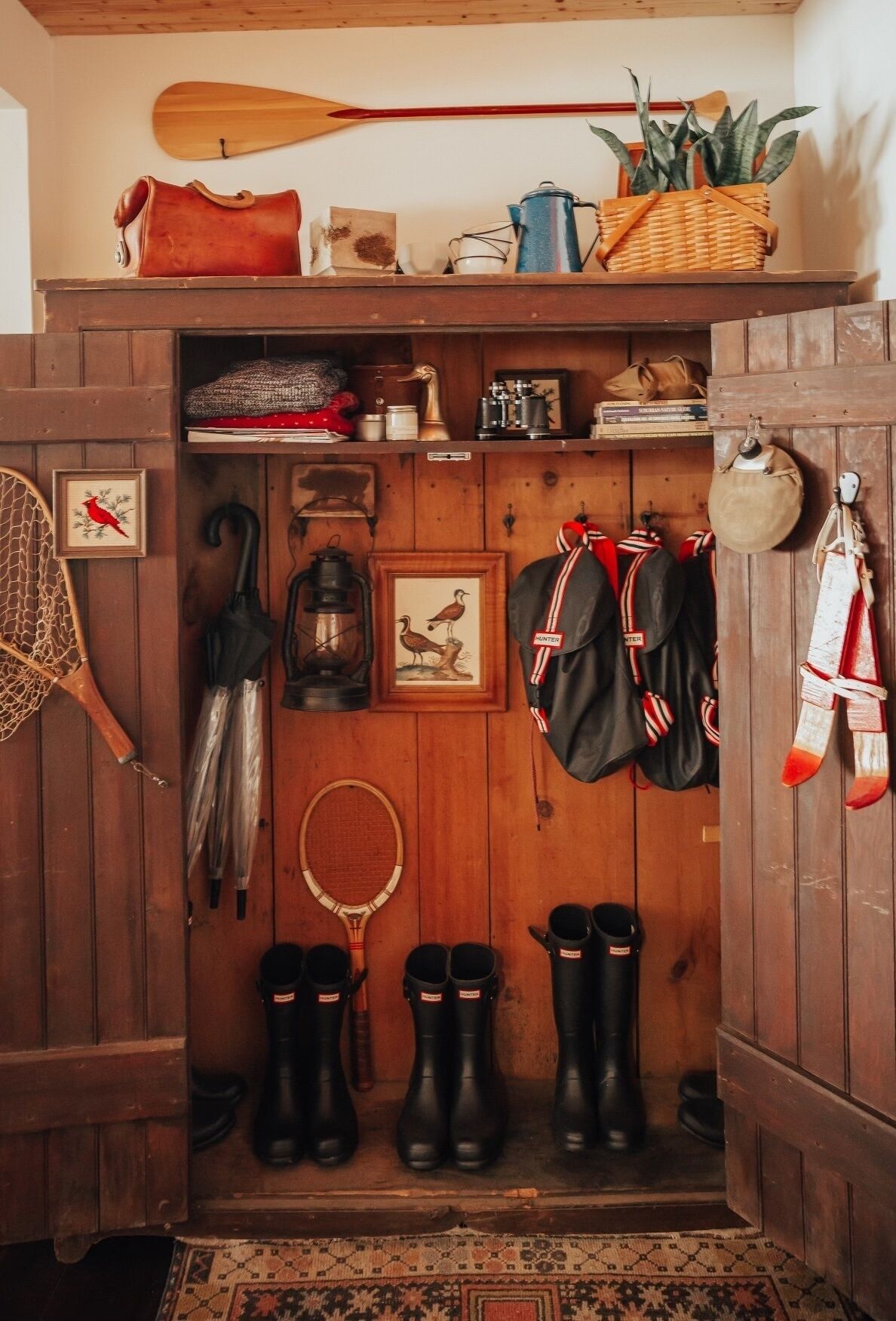 A rustic wooden interior with a shelf holding a paddle, a basket, and a plant. Below, there are boots, a framed picture, and a wooden door.