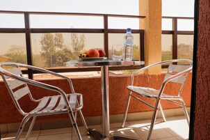 Un balcón con sillas de metal, una mesa con frutas y una botella de agua, y vistas a árboles y edificios.