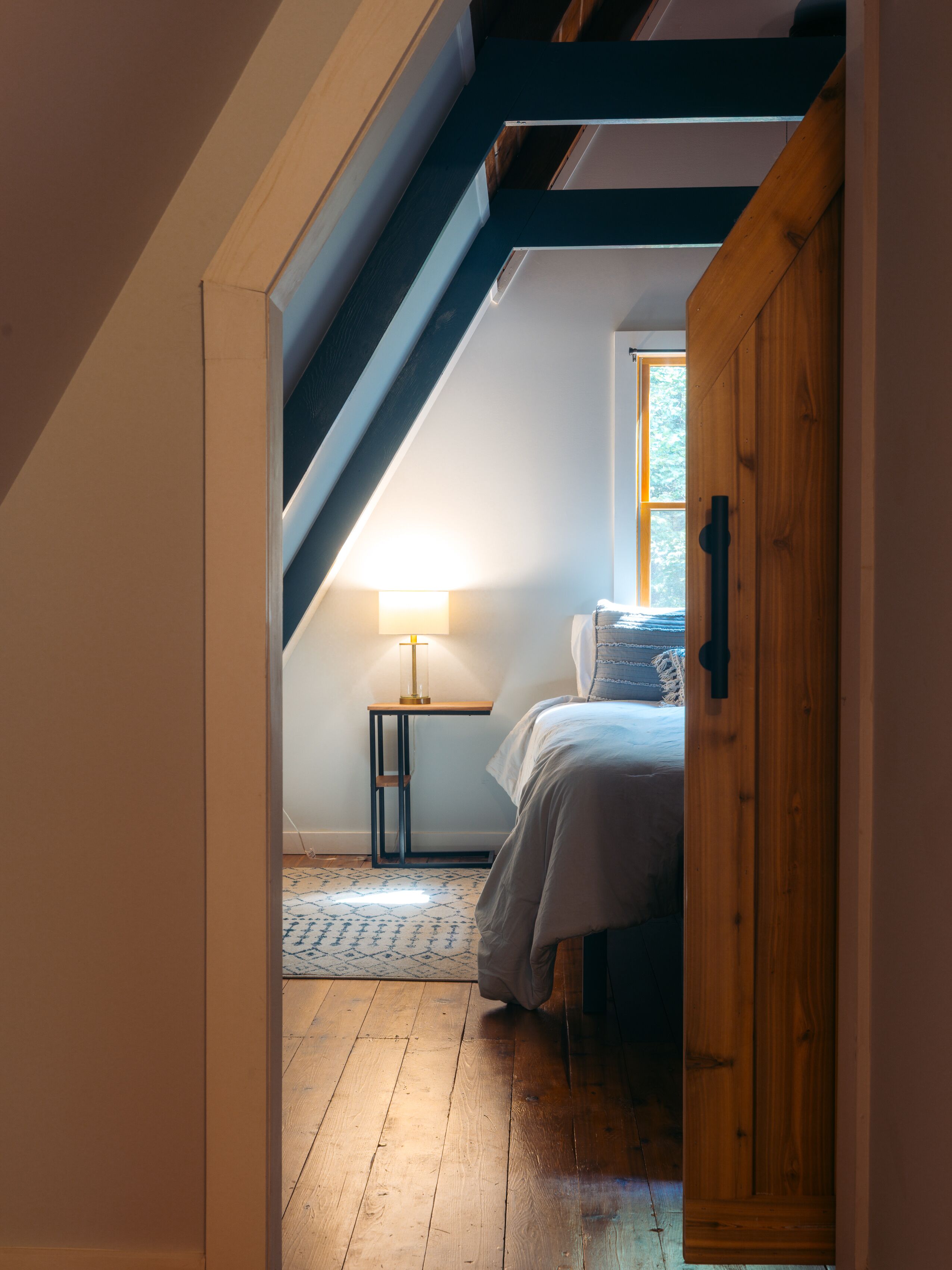 Un dormitorio con techo inclinado, una cama, una mesita de noche con lámpara y una ventana.