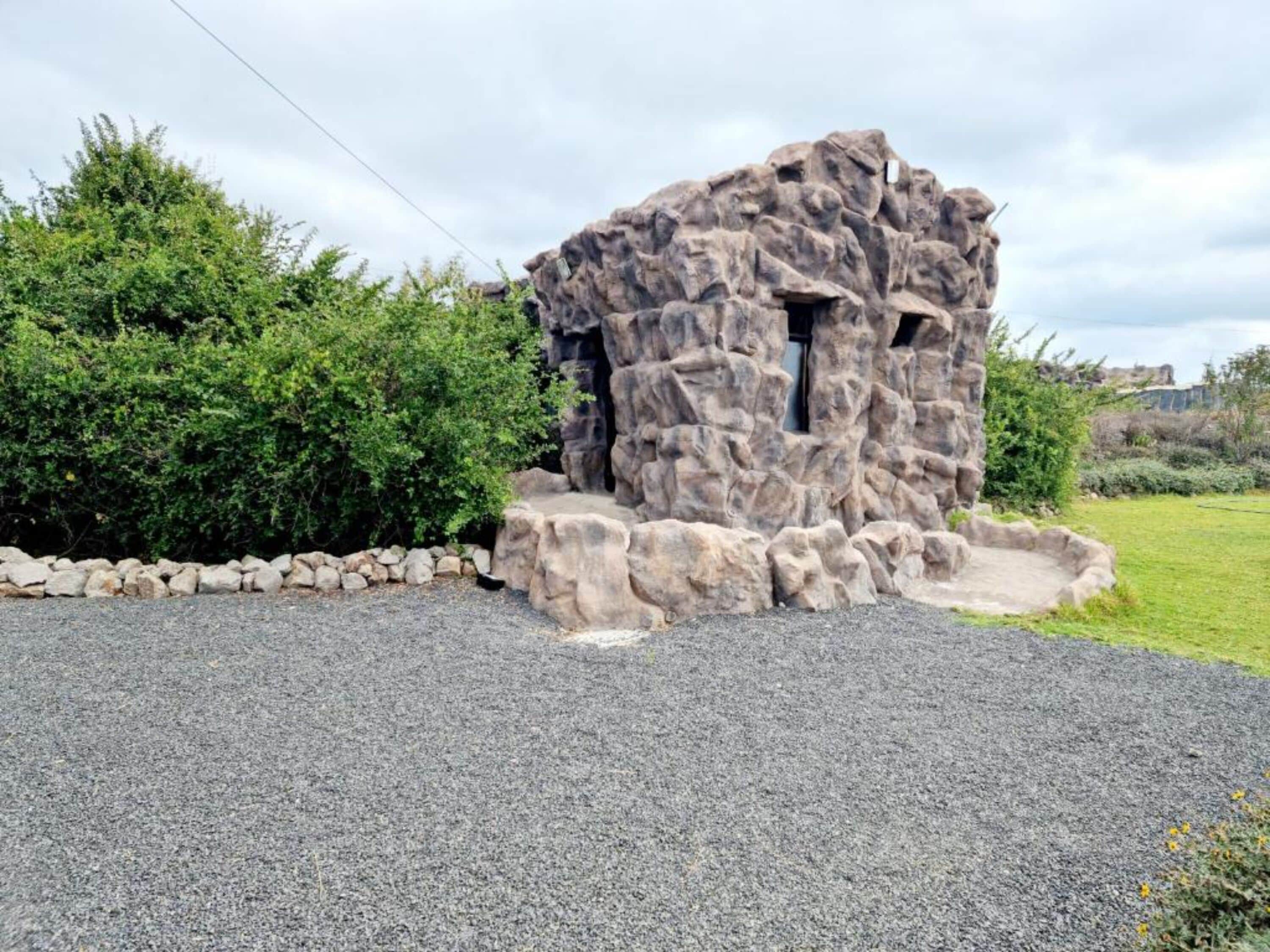 A rock structure with a small opening, surrounded by a stone wall and greenery.