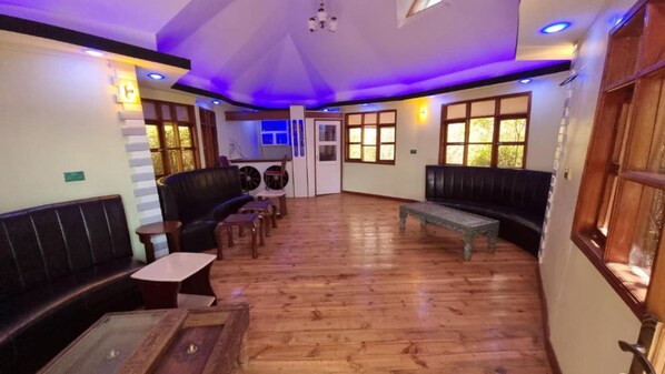 Amplia habitación con piso de madera, un sofá de cuero negro, una mesa de centro de madera y una pequeña mesa con sillas. Cuenta con ventanas de madera y un techo con iluminación empotrada.
