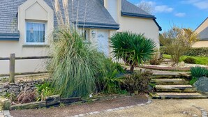 Une maison avec un toit en pente, un mur en pierre et un jardin où poussent différentes plantes, ainsi qu’un escalier en bois.