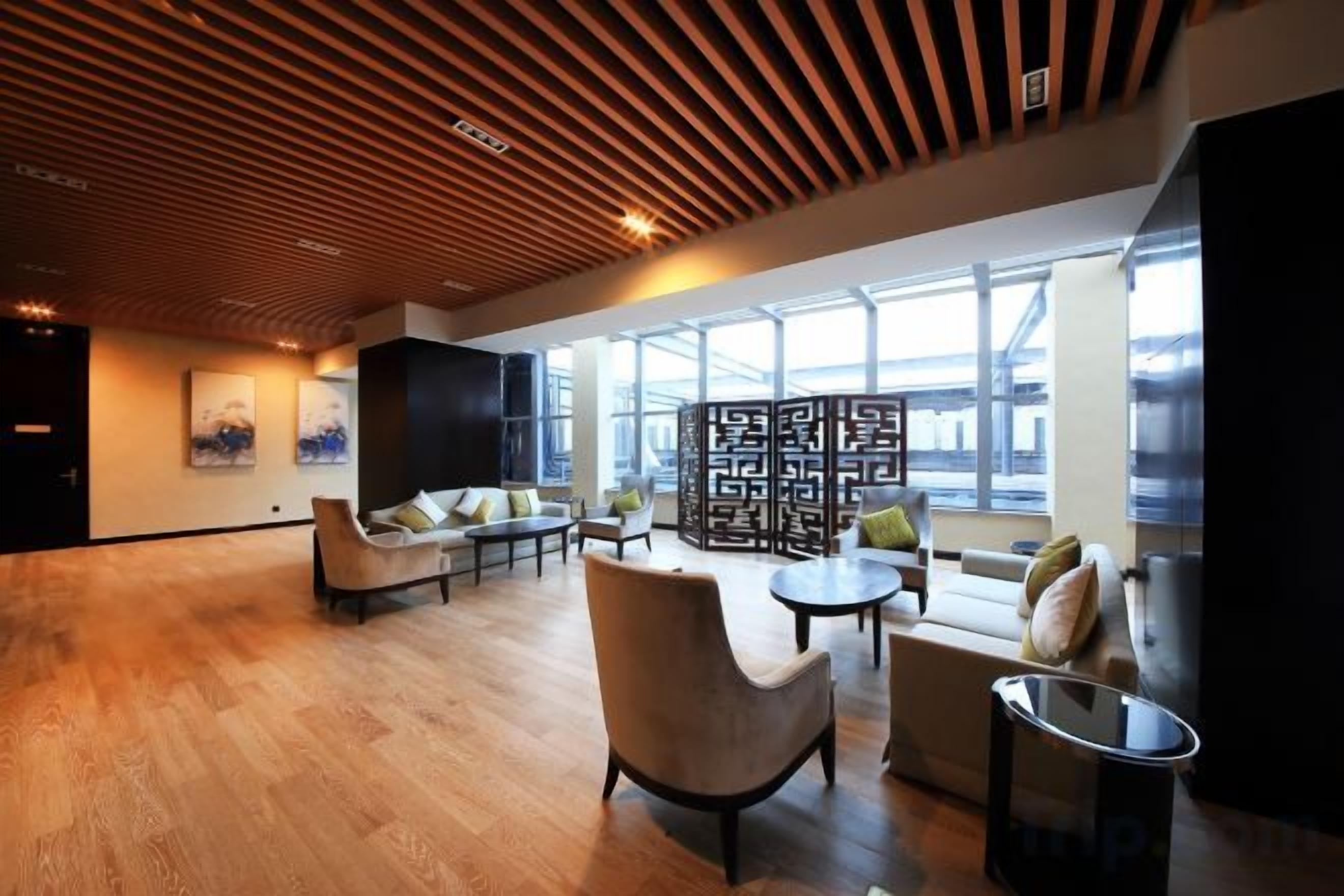 A spacious lobby with wooden flooring, comfortable seating, a large window, and a modern ceiling design.