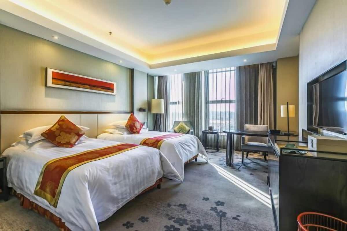 A hotel room with two beds, a desk, a chair, a TV, and a large window.