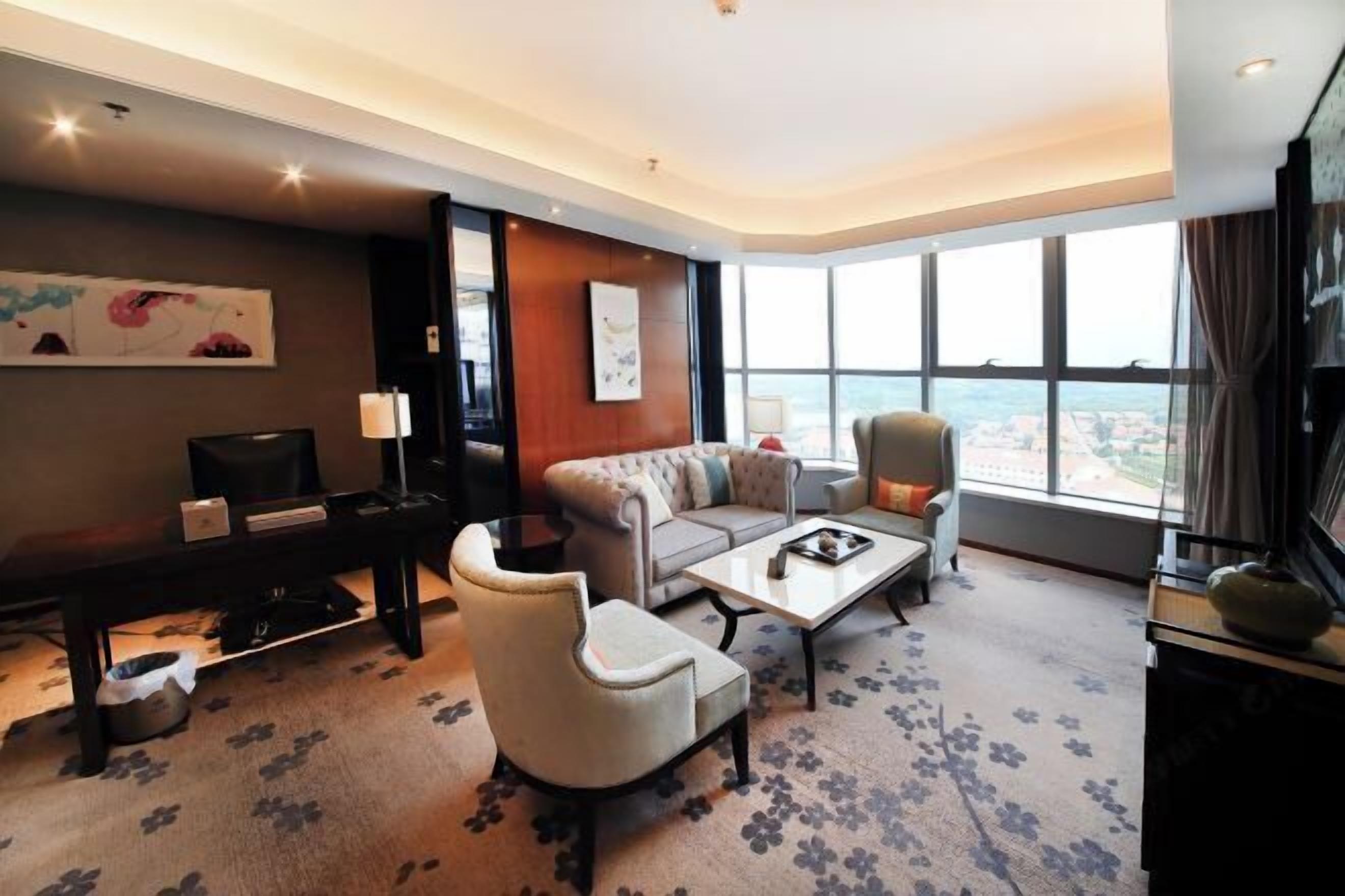 A hotel room with a sofa, armchair, coffee table, and a desk with a lamp.