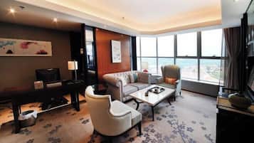 A hotel room with a sofa, armchair, coffee table, and a desk with a lamp.