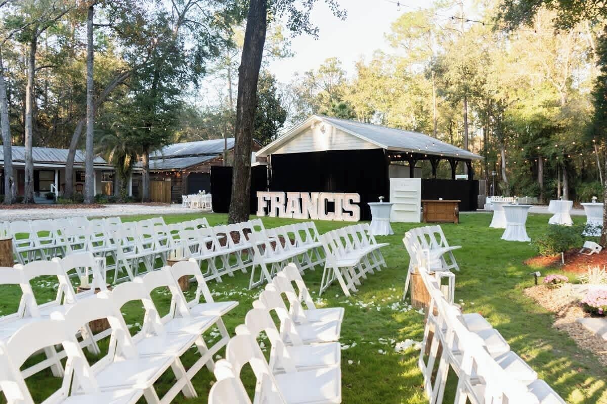 An outdoor seating area with white chairs arranged in rows, a covered structure labeled 'FRANCIS,' and a backdrop of trees.