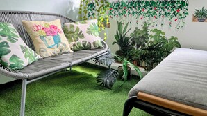 Un canapé en osier avec un coussin, une petite table et un lit dans une pièce dont le sol est recouvert d’un gazon synthétique.