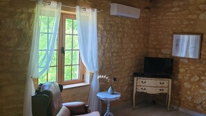 Una habitación con paredes de piedra, un televisor sobre un soporte de madera, una mesita con un jarrón y una ventana con cortinas.