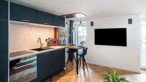 Et moderne køkken med mørke skabe, en træbordplade og en indbygget ovn. Der er et spiseområde med to barstole og et bord, samt et stort fladskærms-tv ophængt på væggen.