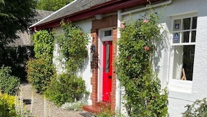 A white cottage with a red door, surrounded by greenery and a gravel path. - Gorgeous Comrie Cottage, hot tub & Mountain Views (Crieff)