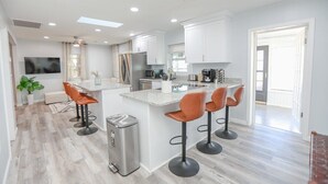 Uma cozinha moderna com armários brancos, uma ilha central com banquetas, uma lata de lixo de aço inoxidável e uma grande janela que proporciona iluminação natural.