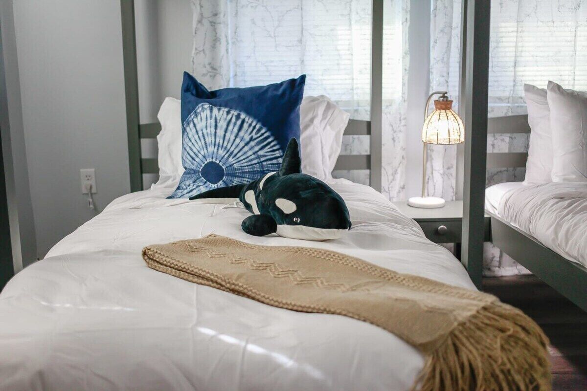 Un lit bien fait, avec une couette blanche, un oreiller bleu et blanc, un jouet en peluche vert et un plaid beige.