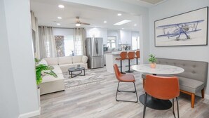 Uma sala de estar moderna com sofá, mesa de centro e cadeiras laranja. Uma área de jantar com mesa redonda e cadeiras é visível. Uma cozinha com frigorífico e fogão pode ser vista ao fundo.