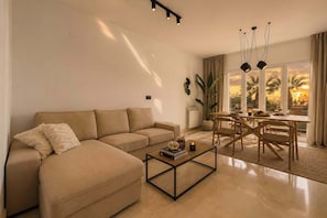 A living room with a sofa, coffee table, dining table, chairs, and a view of palm trees through large windows.