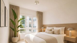 A bedroom with a large bed, two bedside lamps, a potted plant, and a window with curtains.