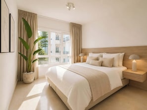 A bedroom with a large bed, two bedside lamps, a potted plant, and a window with curtains.