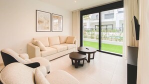 A modern living room with a sofa, two armchairs, a coffee table, and a TV stand. There is a large window with a view of buildings and greenery.
