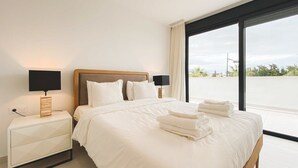 A modern hotel room with a large bed, a bedside table with a lamp, and a view of the outdoors through a large window.