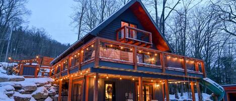 A two-story log cabin with a balcony, surrounded by snow and trees.