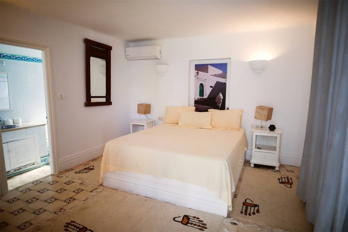 Ein Schlafzimmer mit einem Bett, einem Nachttisch, einer Klimaanlage, einem Poster an der Wand und einem durch eine offene Tür einsehbaren Badezimmer.