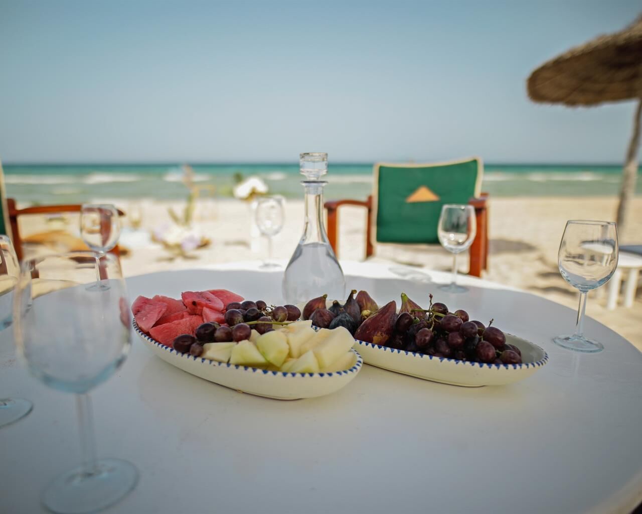 Ein Tisch mit Obstplatten und einer Flasche, auf einem Strand mit Stühlen und Blick aufs Meer.
