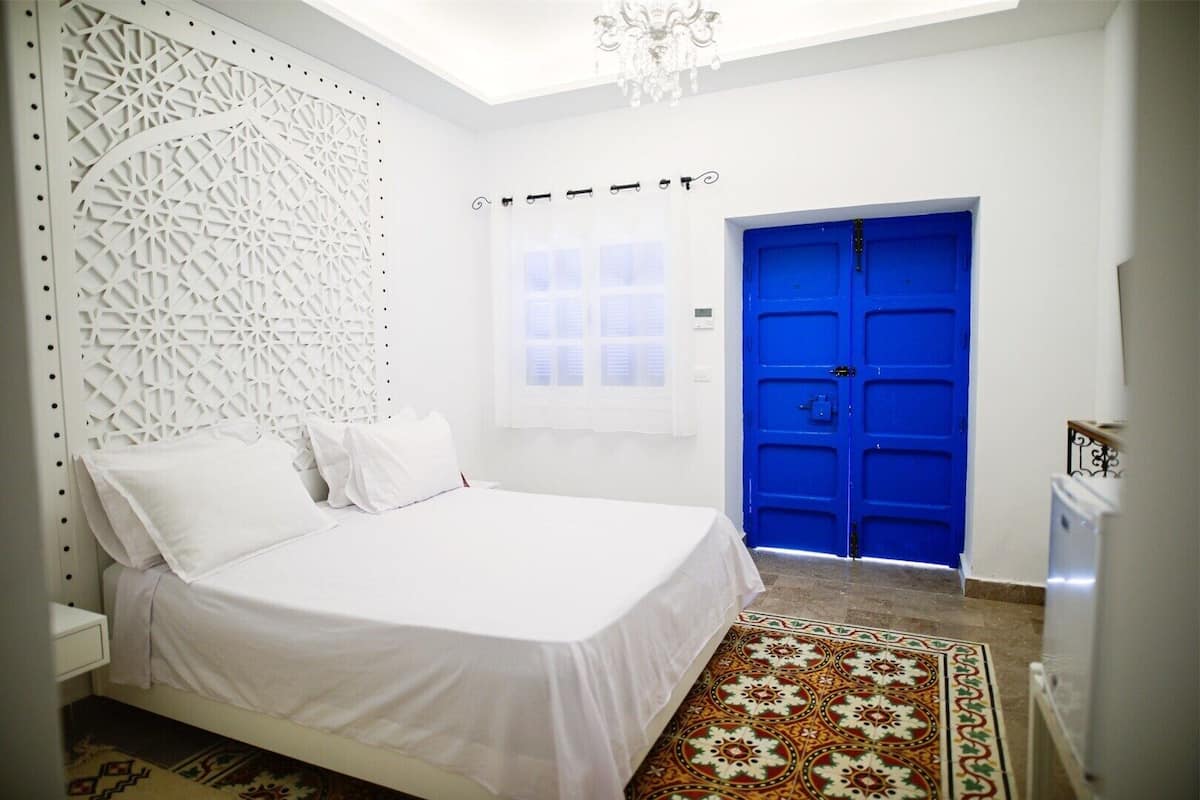 Ein Schlafzimmer mit Bett, weißen Wänden, einer blauen Tür und einem gemusterten Teppich.