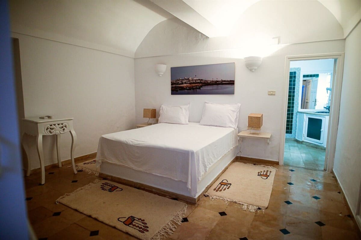 Ein Schlafzimmer mit Bett, Nachttisch, kleinem Tisch, einem Bild an der Wand und Blick ins Freie.