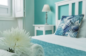 A neatly made bed with a white duvet, a blue patterned bedspread, and a blue leaf-patterned pillow. A white bedside table with a lamp and a white towel is visible. - Lalitana Townhouse (Lagos)