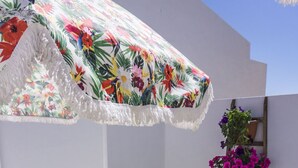 A small outdoor patio with a white table and two chairs, a floral umbrella, and potted plants on a white wall. - Lalitana Townhouse (Lagos)