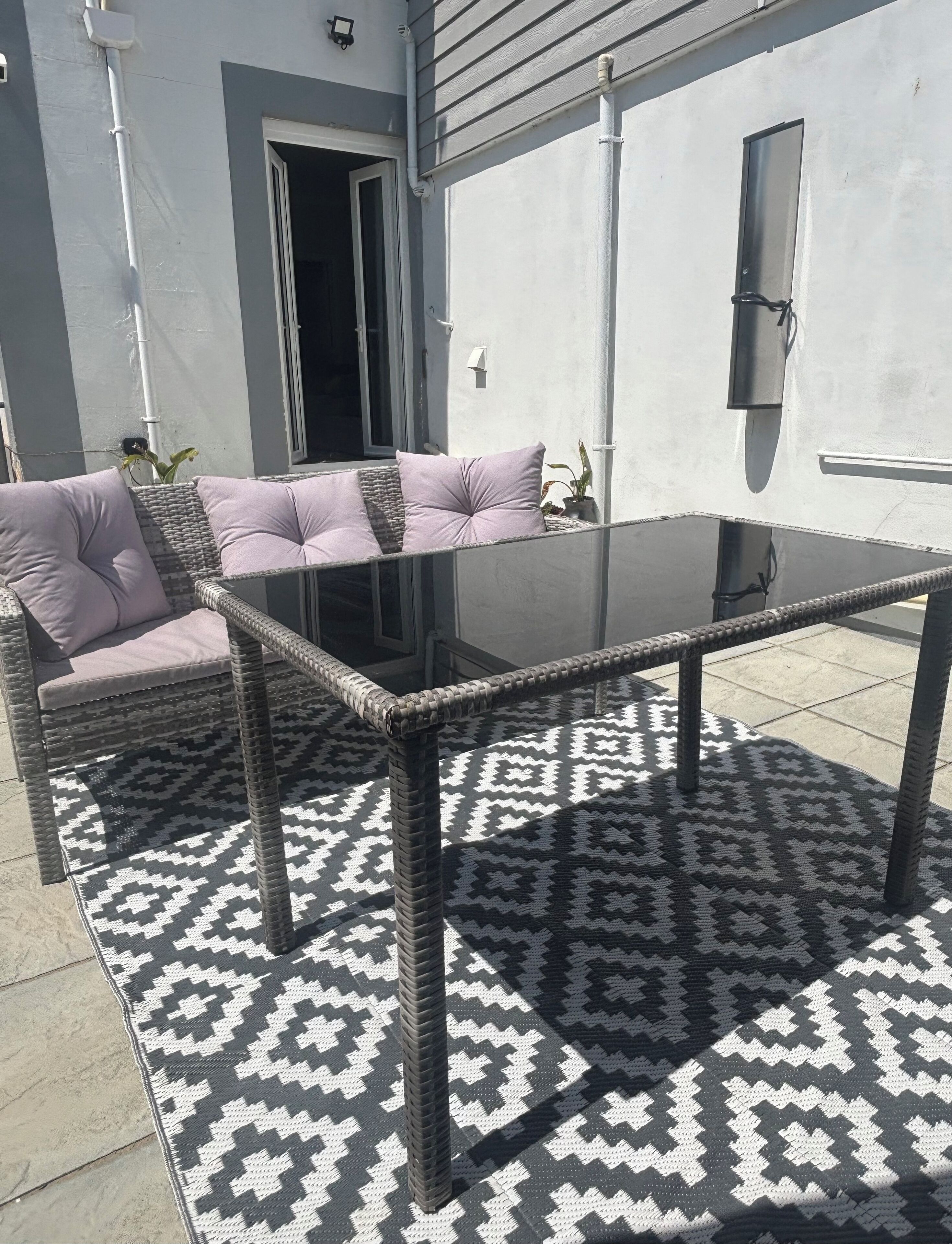 Une terrasse extérieure avec une table à plateau en verre, des meubles en osier et des tapis à motifs.