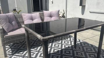 Une terrasse extérieure avec une table à plateau en verre, des meubles en osier et des tapis à motifs.