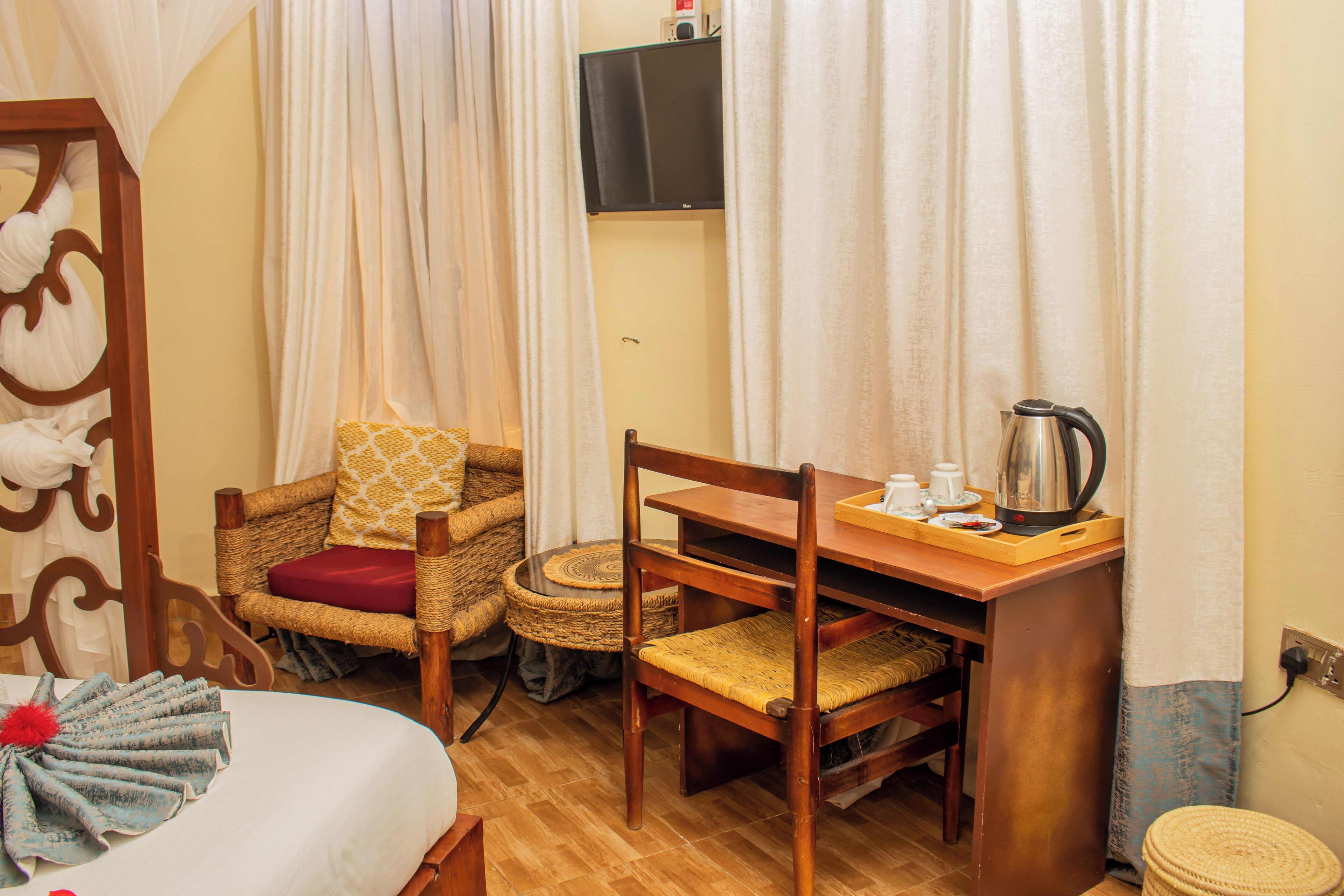 A cozy room with a bed, a wicker chair, a wooden table with a kettle, and a television on the wall.