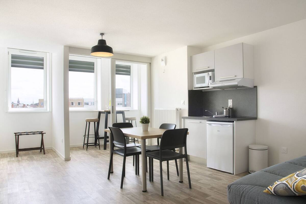 Eine kompakte Küche mit Mikrowelle, kleinem Kühlschrank und einer schwarzen Arbeitsplatte. Ein Essbereich mit Tisch und Stühlen sowie ein graues Sofa mit einem gemusterten Kissen.