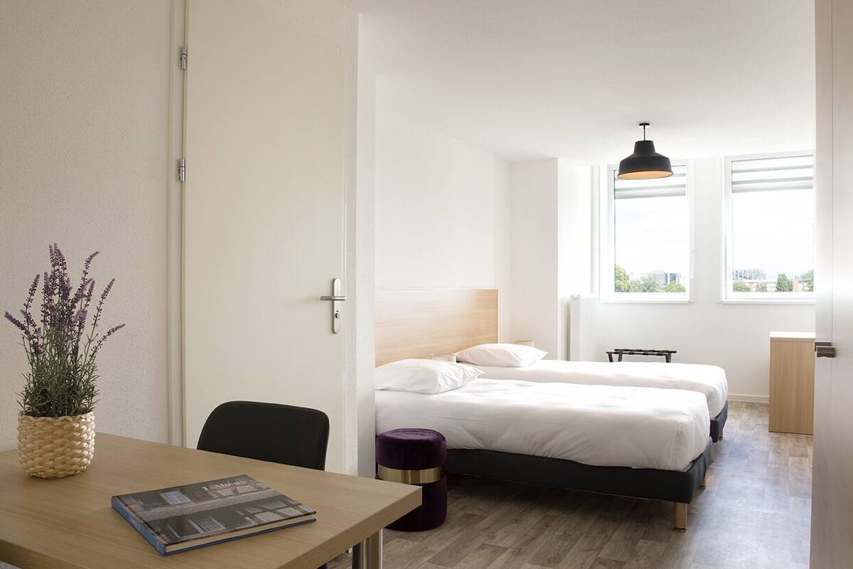 Ein Hotelzimmer mit zwei Betten, einem Schreibtisch mit Stuhl, einem kleinen Tisch mit Pflanze und einem Fenster mit Blick auf Gebäude.