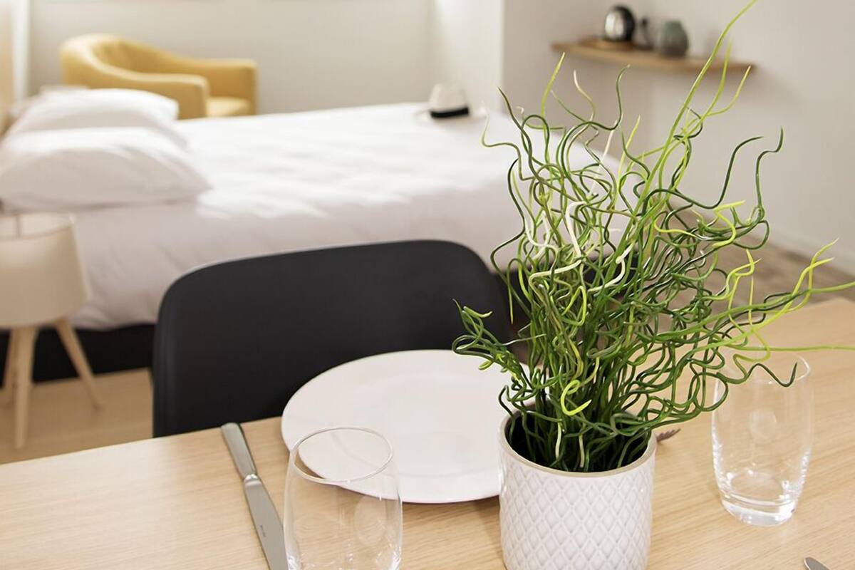 Ein ordentlich eingerichtetes Hotelzimmer mit einem Bett, einem Nachttisch mit einem Glas Wasser, einer Topfpflanze und einem an der Wand montierten Fernseher.